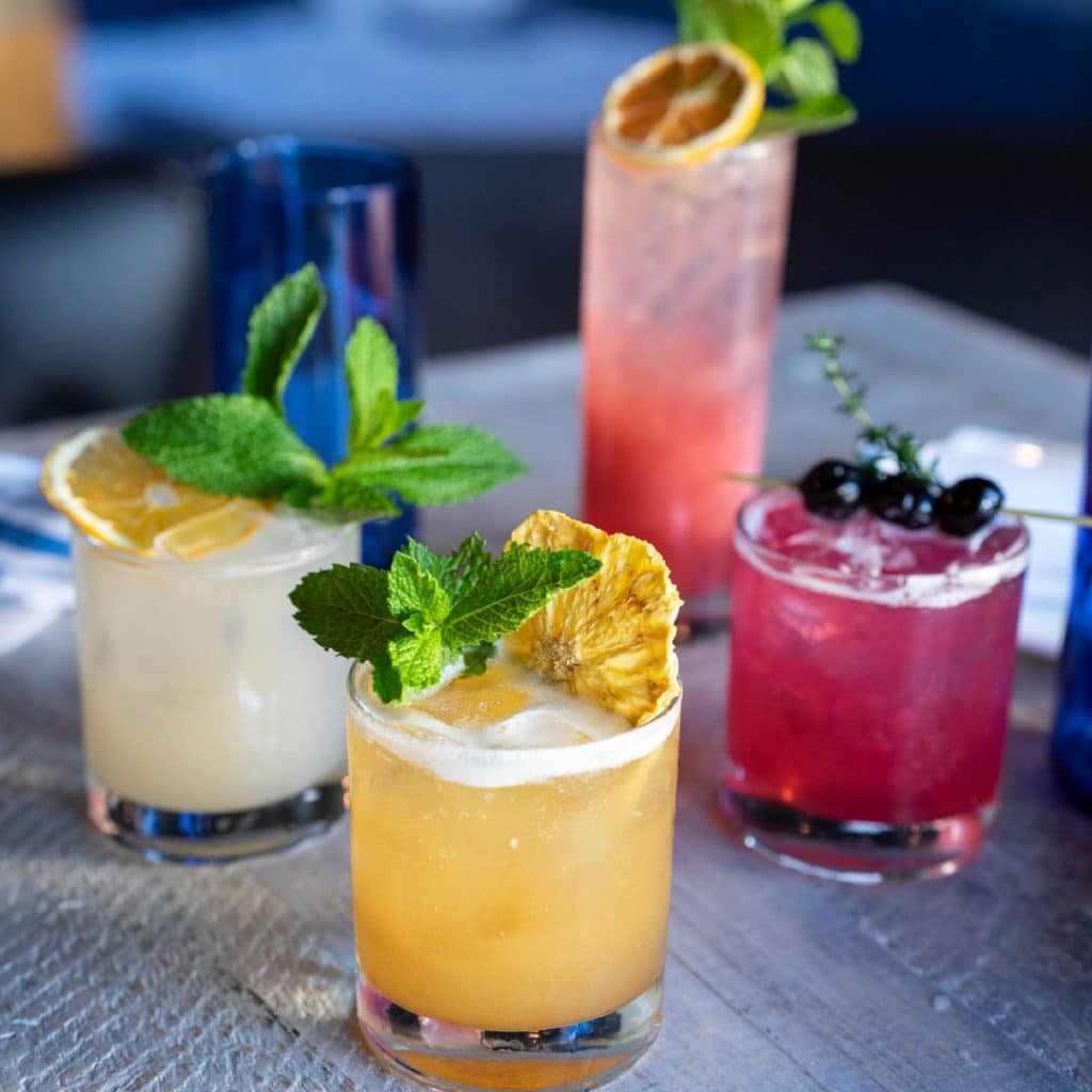 Close-up of a yellow-orange cocktail in the foreground, garnished with a mint sprig and a dried pineapple wheel, with white, pink, and red drinks in the background on a metallic table.
