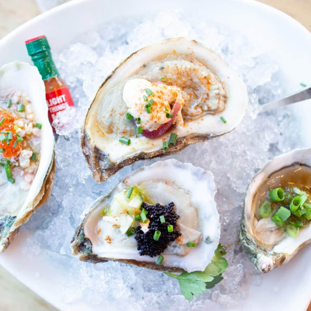 Platter of raw oysters on ice with assorted toppings and a hot sauce bottle in the background.