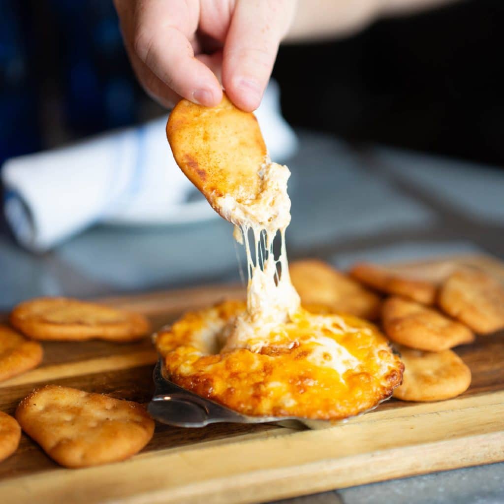 Hand lifting a crisp cracker to dip into gooey cheese fondue on a wooden board.